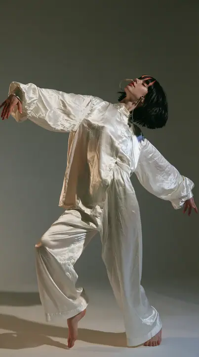 full-length shot, a Caucasian American woman in her late twenties with pale skin and a glossy black blunt bob with heavy straight bangs and one clay-orange streak, a thin headset microphone, in oversized white…