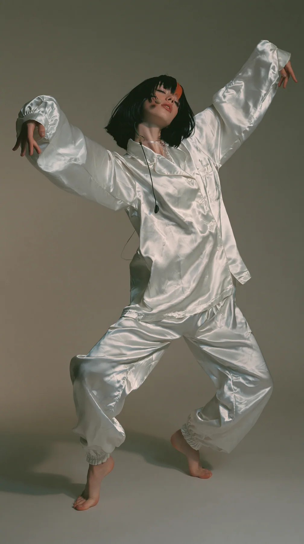 full-length shot, a Caucasian American woman in her late twenties with pale skin and a glossy black blunt bob with heavy straight bangs and one clay-orange streak, a thin headset microphone, in oversized white…