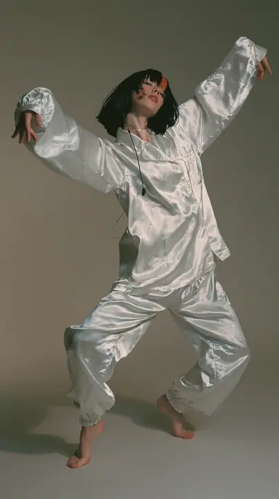 full-length shot, a Caucasian American woman in her late twenties with pale skin and a glossy black blunt bob with heavy straight bangs and one clay-orange streak, a thin headset microphone, in oversized white…