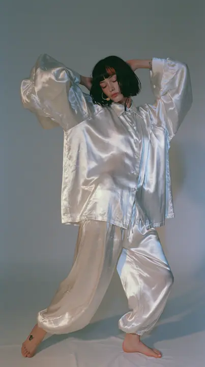 full-length shot, a Caucasian American woman in her late twenties with pale skin and a glossy black blunt bob with heavy straight bangs and one clay-orange streak, a thin headset microphone, in oversized white…