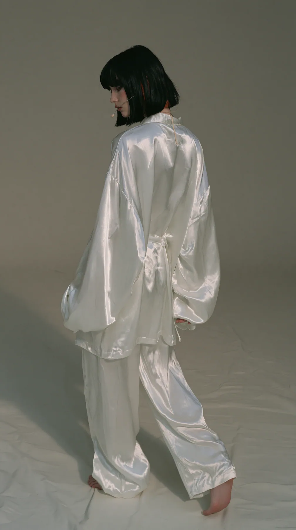 full-length shot from behind, a Caucasian American woman in her late twenties with pale skin and a glossy black blunt bob with heavy straight bangs and one clay-orange streak, a thin headset microphone, in oversized…