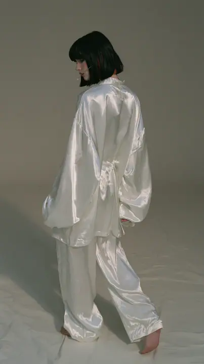 full-length shot from behind, a Caucasian American woman in her late twenties with pale skin and a glossy black blunt bob with heavy straight bangs and one clay-orange streak, a thin headset microphone, in oversized…