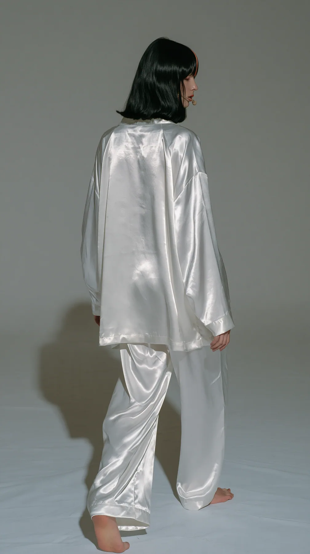 full-length shot from behind, a Caucasian American woman in her late twenties with pale skin and a glossy black blunt bob with heavy straight bangs and one clay-orange streak, a thin headset microphone, in oversized…