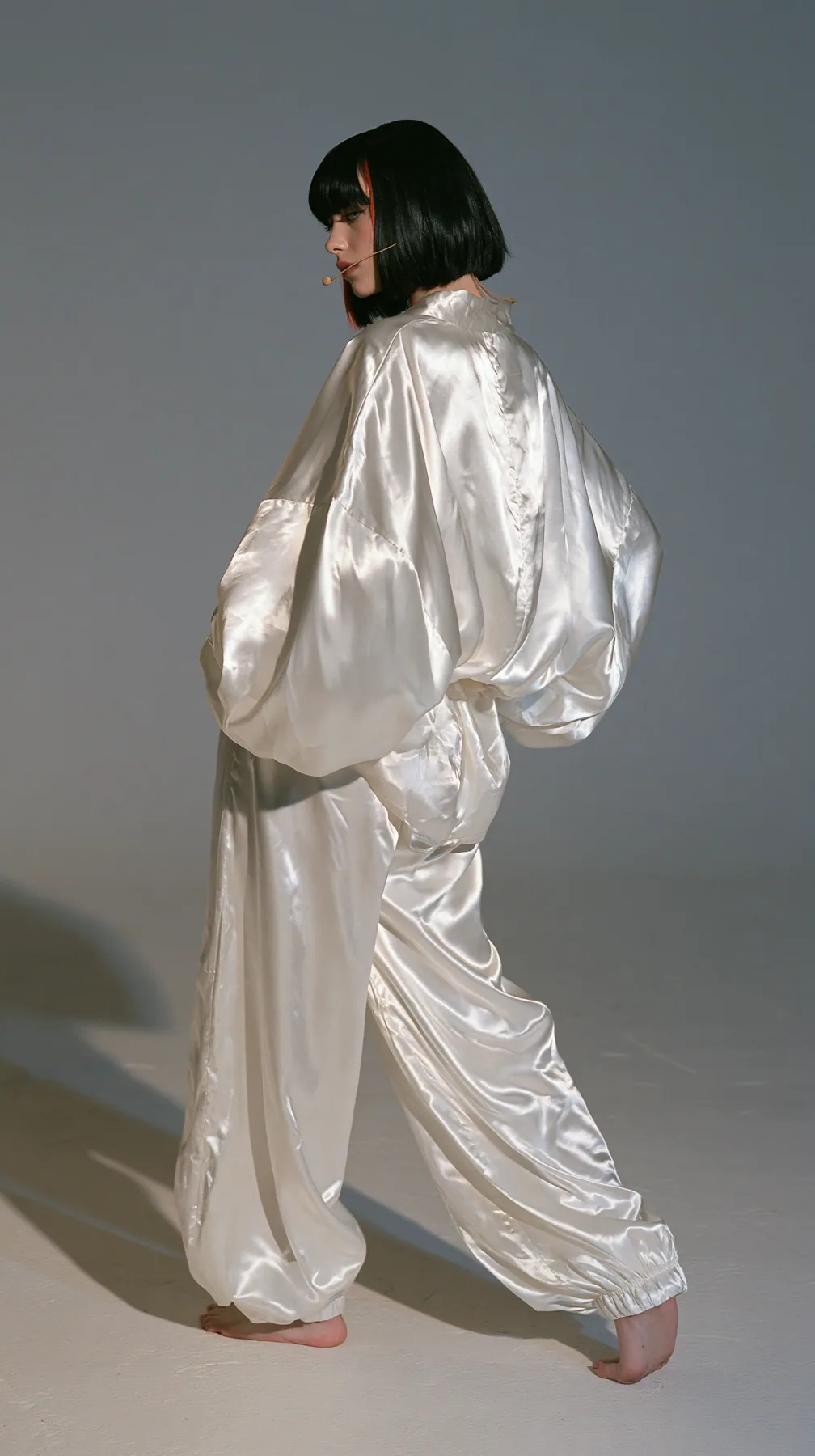 full-length shot from behind, a Caucasian American woman in her late twenties with pale skin and a glossy black blunt bob with heavy straight bangs and one clay-orange streak, a thin headset microphone, in oversized…
