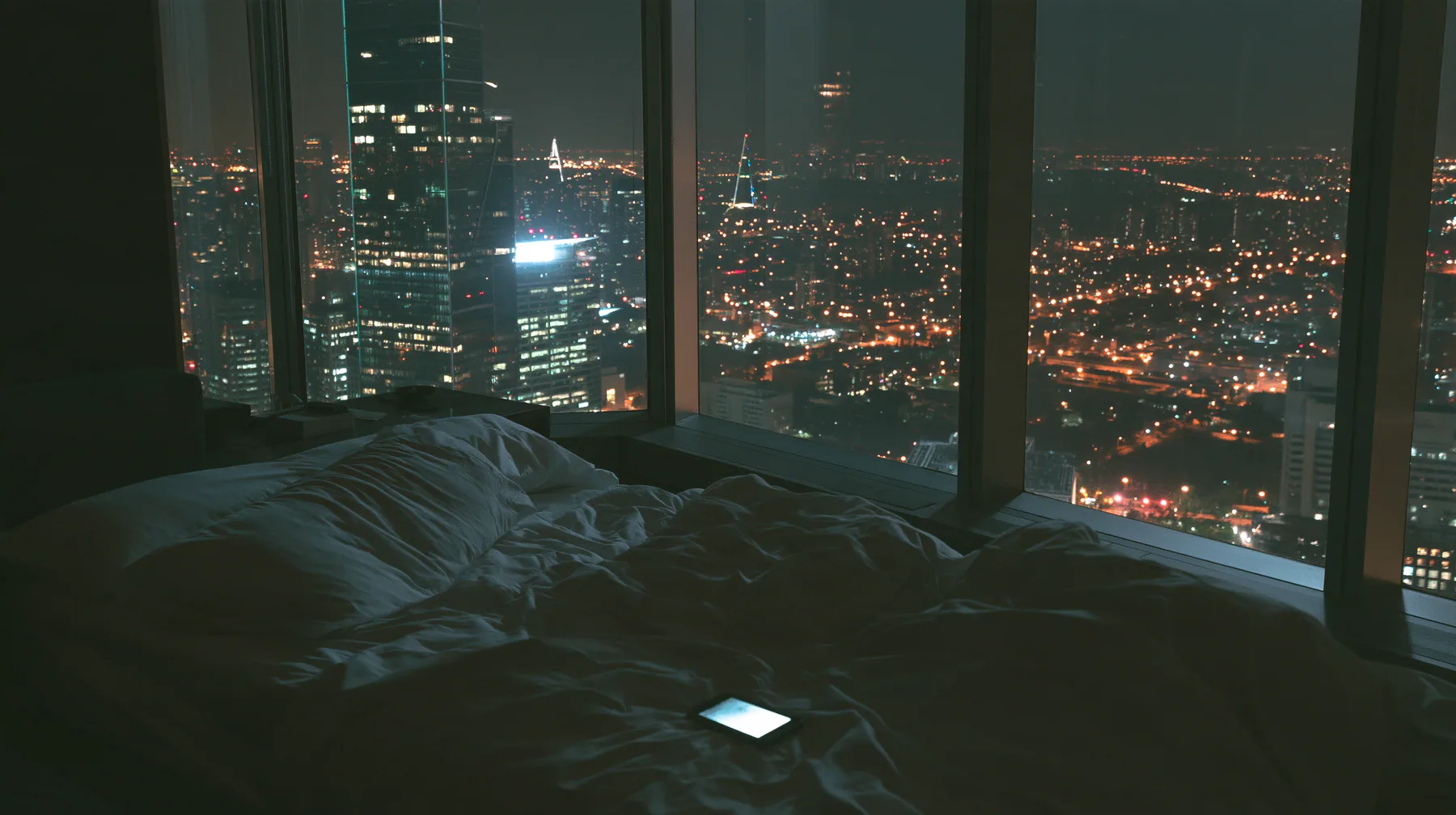 a dark minimal high-rise bedroom at night, a smartphone lying face down on rumpled white sheets, a floor-to-ceiling window of glittering city lights behind, no people, deep shadows, cinematic, no text, no letters, no…