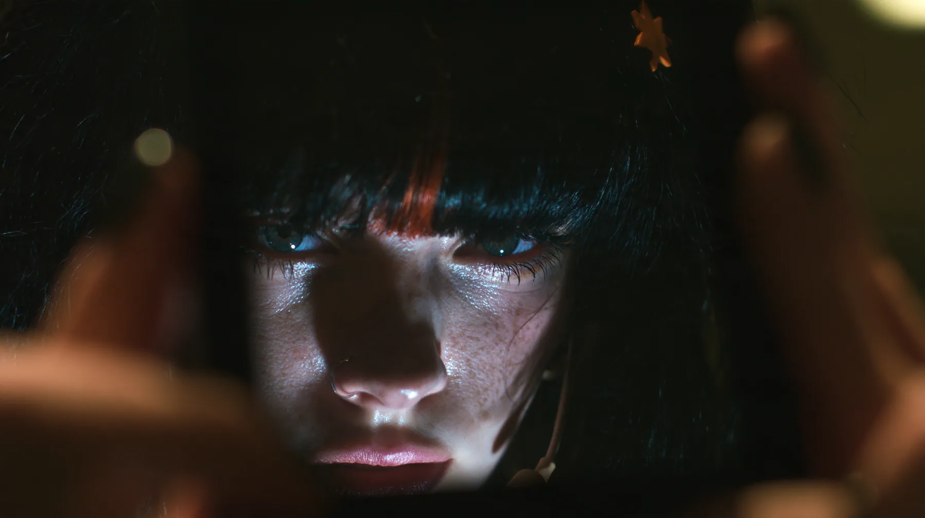 extreme close-up of a dead black phone screen held in two hands in near darkness, faintly reflecting the face of a 28-year-old Caucasian American woman with a grown-up angular face, defined cheekbones and a strong…