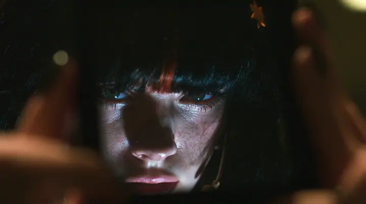 extreme close-up of a dead black phone screen held in two hands in near darkness, faintly reflecting the face of a 28-year-old Caucasian American woman with a grown-up angular face, defined cheekbones and a strong…