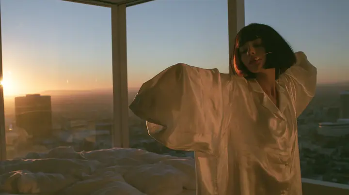 wide shot of a minimal high-rise bedroom at dawn, golden sunrise through a floor-to-ceiling window, a Caucasian American woman in her late twenties with pale skin and a glossy black blunt bob with heavy straight…