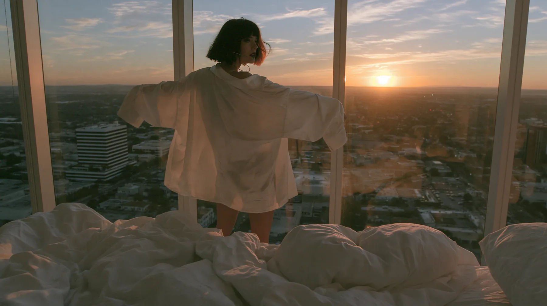 wide shot of a minimal high-rise bedroom at dawn, golden sunrise through a floor-to-ceiling window, a Caucasian American woman in her late twenties with pale skin and a glossy black blunt bob with heavy straight…