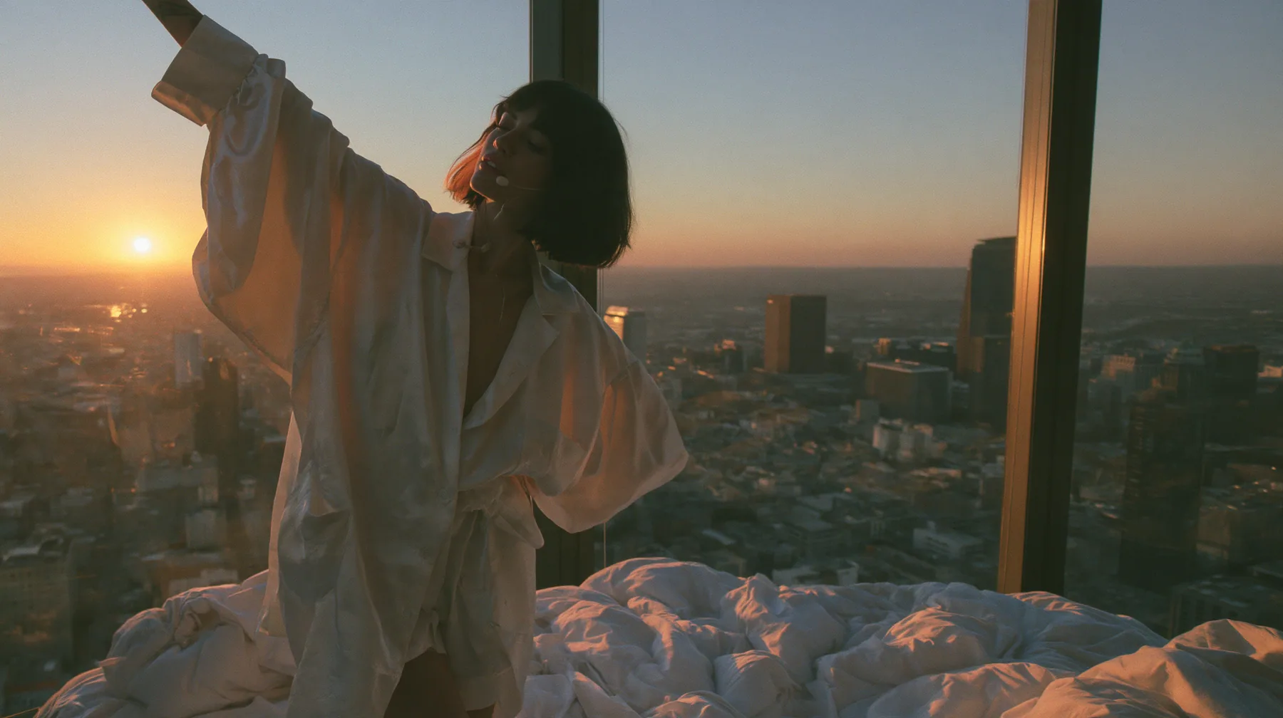 wide shot of a minimal high-rise bedroom at dawn, golden sunrise through a floor-to-ceiling window, a Caucasian American woman in her late twenties with pale skin and a glossy black blunt bob with heavy straight…