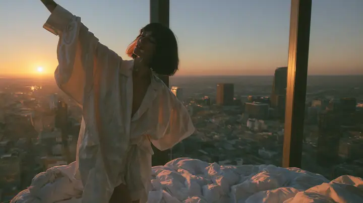 wide shot of a minimal high-rise bedroom at dawn, golden sunrise through a floor-to-ceiling window, a Caucasian American woman in her late twenties with pale skin and a glossy black blunt bob with heavy straight…