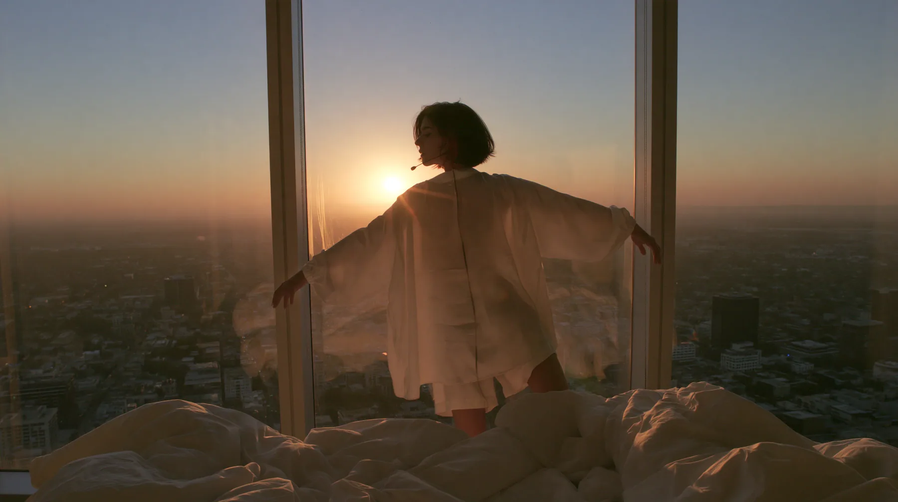 wide shot of a minimal high-rise bedroom at dawn, golden sunrise through a floor-to-ceiling window, a Caucasian American woman in her late twenties with pale skin and a glossy black blunt bob with heavy straight…