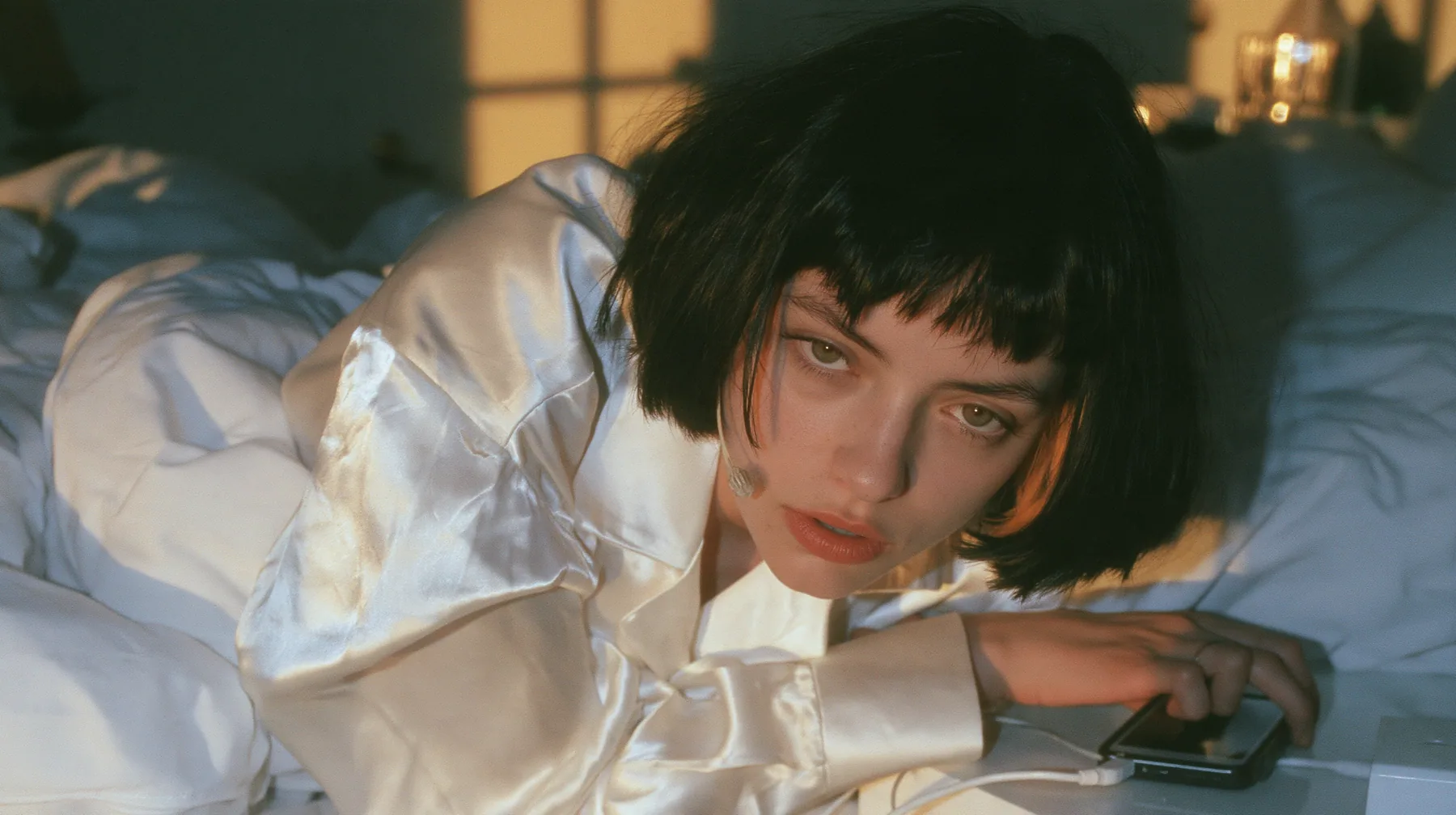 medium shot of a bedside at dawn, a Caucasian American woman in her late twenties with pale skin and a glossy black blunt bob with heavy straight bangs and one clay-orange streak, a thin headset microphone, in…