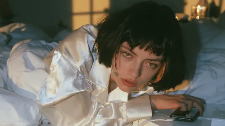 medium shot of a bedside at dawn, a Caucasian American woman in her late twenties with pale skin and a glossy black blunt bob with heavy straight bangs and one clay-orange streak, a thin headset microphone, in…