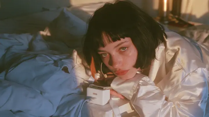 medium shot of a bedside at dawn, a Caucasian American woman in her late twenties with pale skin and a glossy black blunt bob with heavy straight bangs and one clay-orange streak, a thin headset microphone, in…