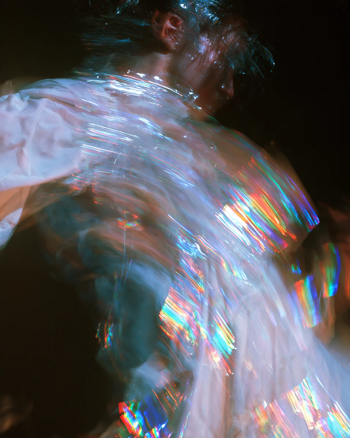 an abstract motion-blurred photograph of a dancing figure dissolving into streaks of iridescent light, black background, no face, no text, no letters, no logos