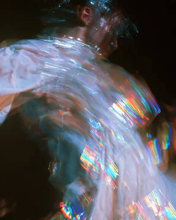 an abstract motion-blurred photograph of a dancing figure dissolving into streaks of iridescent light, black background, no face, no text, no letters, no logos
