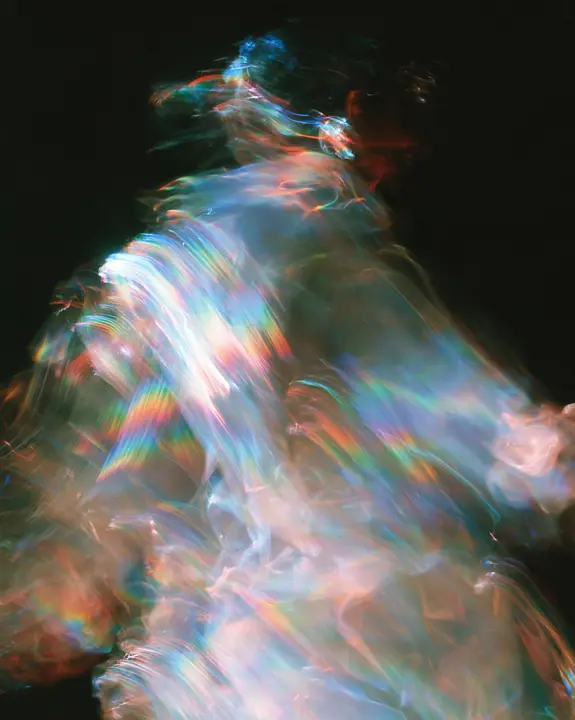 an abstract motion-blurred photograph of a dancing figure dissolving into streaks of iridescent light, black background, no face, no text, no letters, no logos