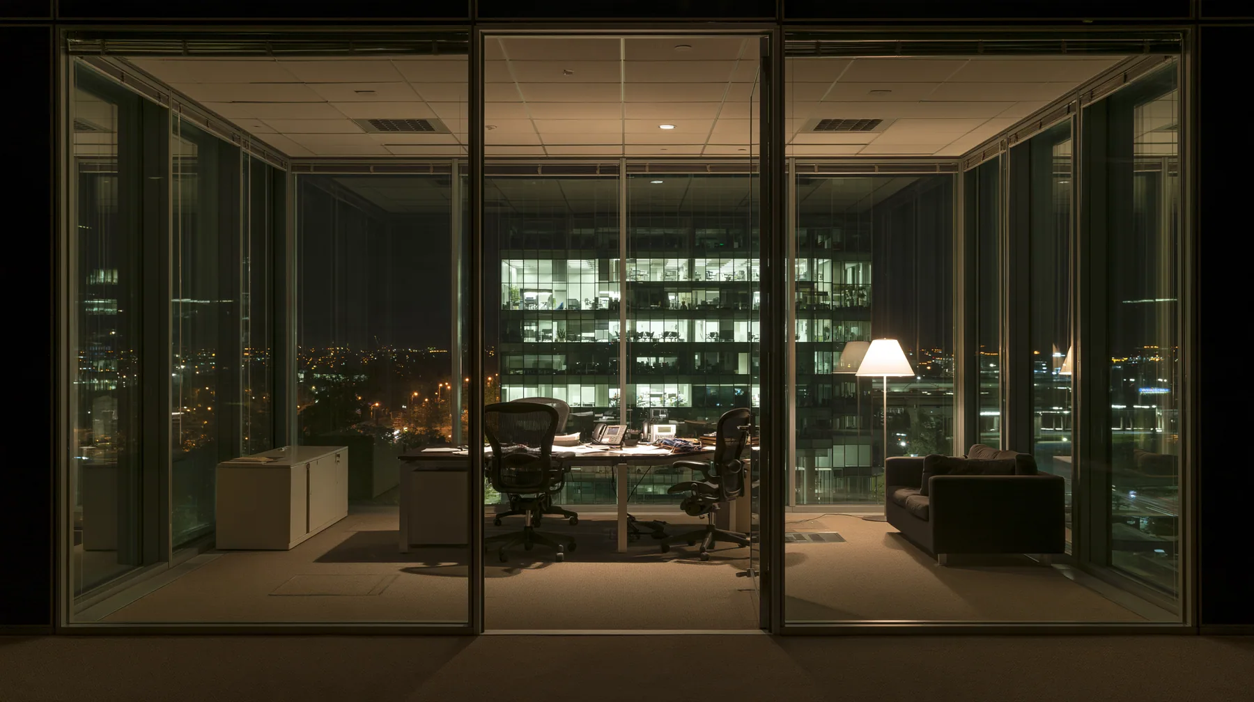 an empty glass-walled office at night, city lights, one lamp left on, wide angle, no text, no letters, no logos