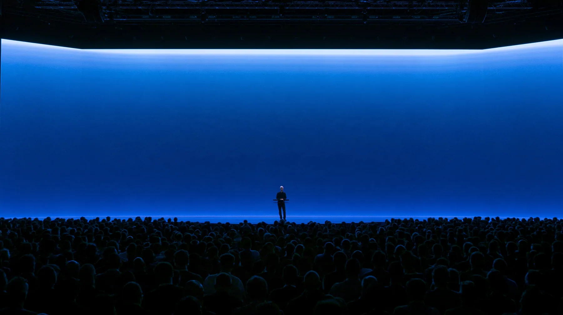 a keynote stage seen from the back of a dark auditorium, one small figure at a podium under a vast glowing blue screen, no text, no letters, no logos