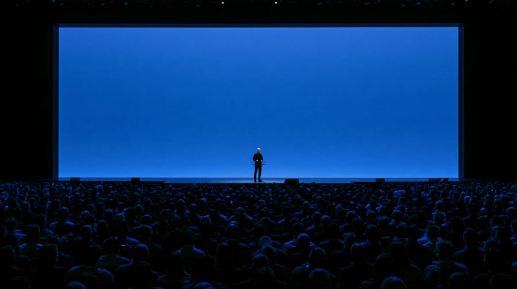 a keynote stage seen from the back of a dark auditorium, one small figure at a podium under a vast glowing blue screen, no text, no letters, no logos
