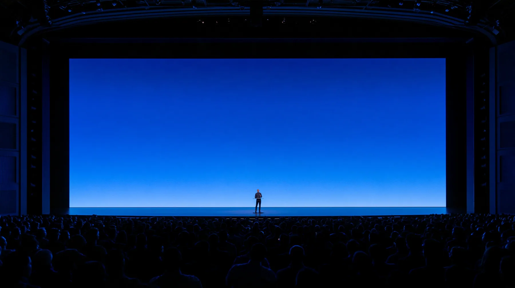 a keynote stage seen from the back of a dark auditorium, one small figure at a podium under a vast glowing blue screen, no text, no letters, no logos