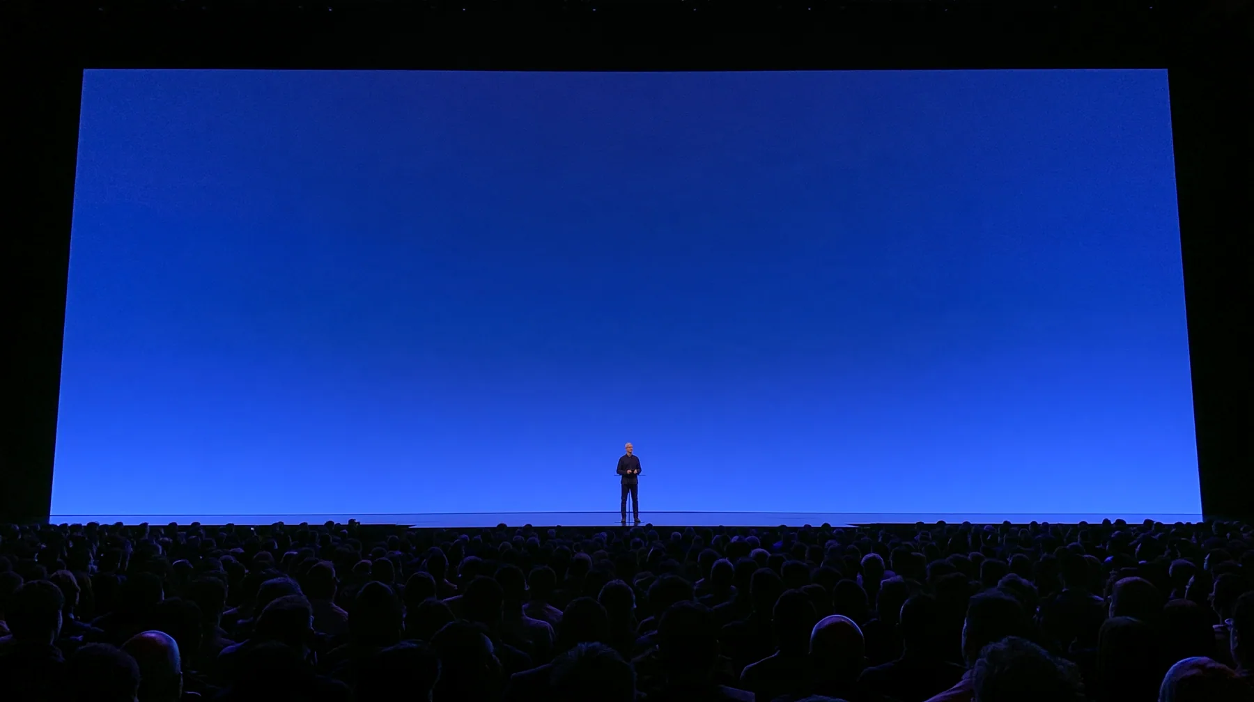 a keynote stage seen from the back of a dark auditorium, one small figure at a podium under a vast glowing blue screen, no text, no letters, no logos