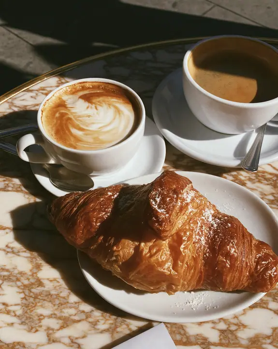 a flaky croissant on a marble cafe table with a cappuccino, morning light, no text, no letters, no logos