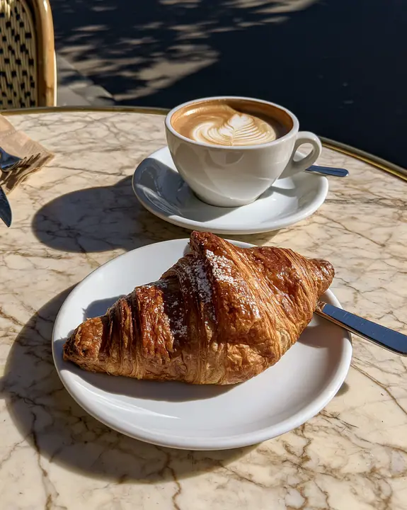 a flaky croissant on a marble cafe table with a cappuccino, morning light, no text, no letters, no logos