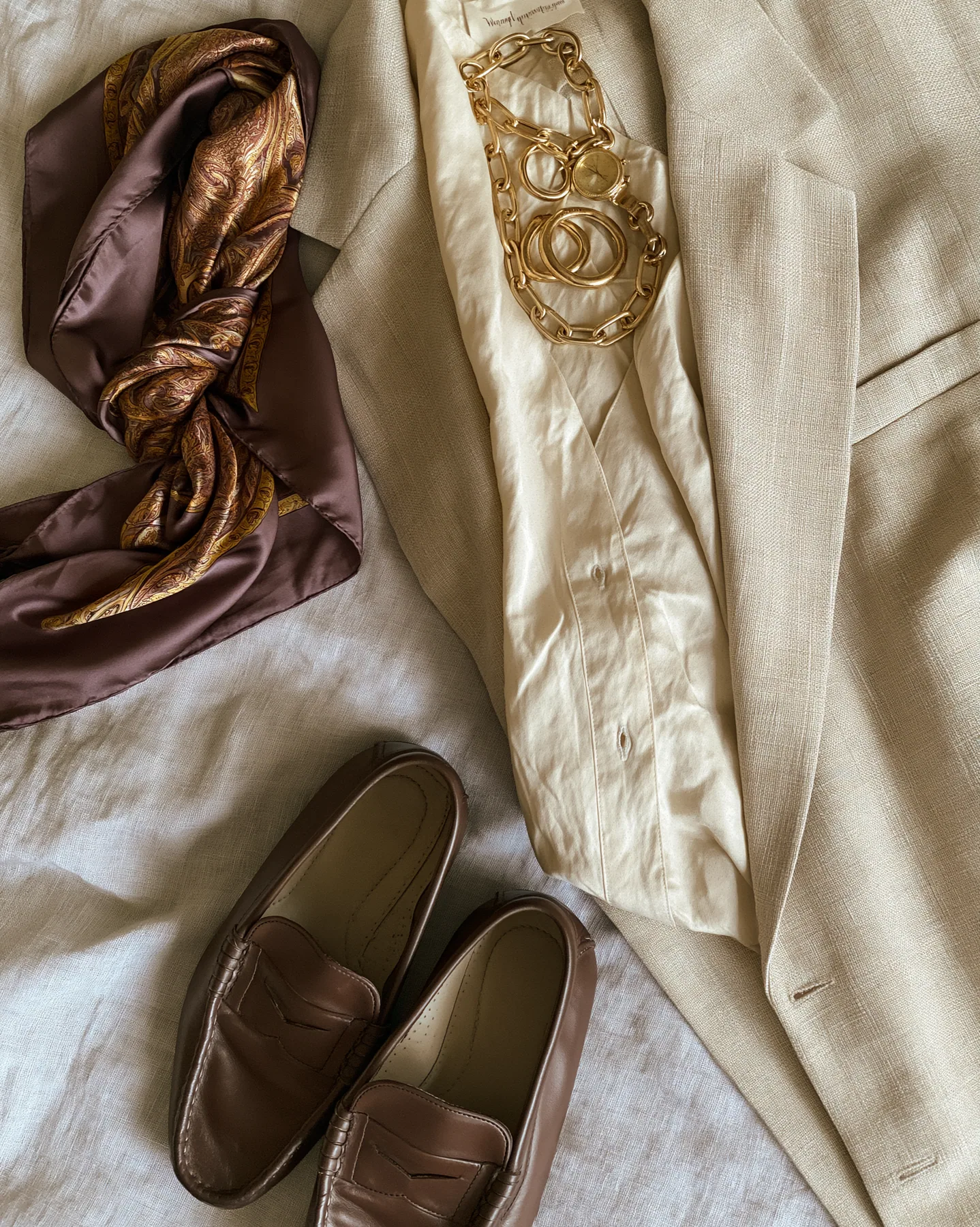 a fashion flat lay of an oversized blazer, silk scarf, loafers and gold jewelry on linen, overhead, no text, no letters, no logos