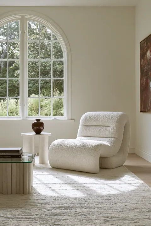 a minimal cream living room with a boucle chair and an arched window, morning light, interior design photograph, no text, no letters, no logos