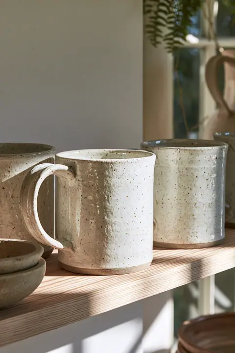 handmade speckled ceramic mugs on a pale wooden shelf, soft light, no text, no letters, no logos