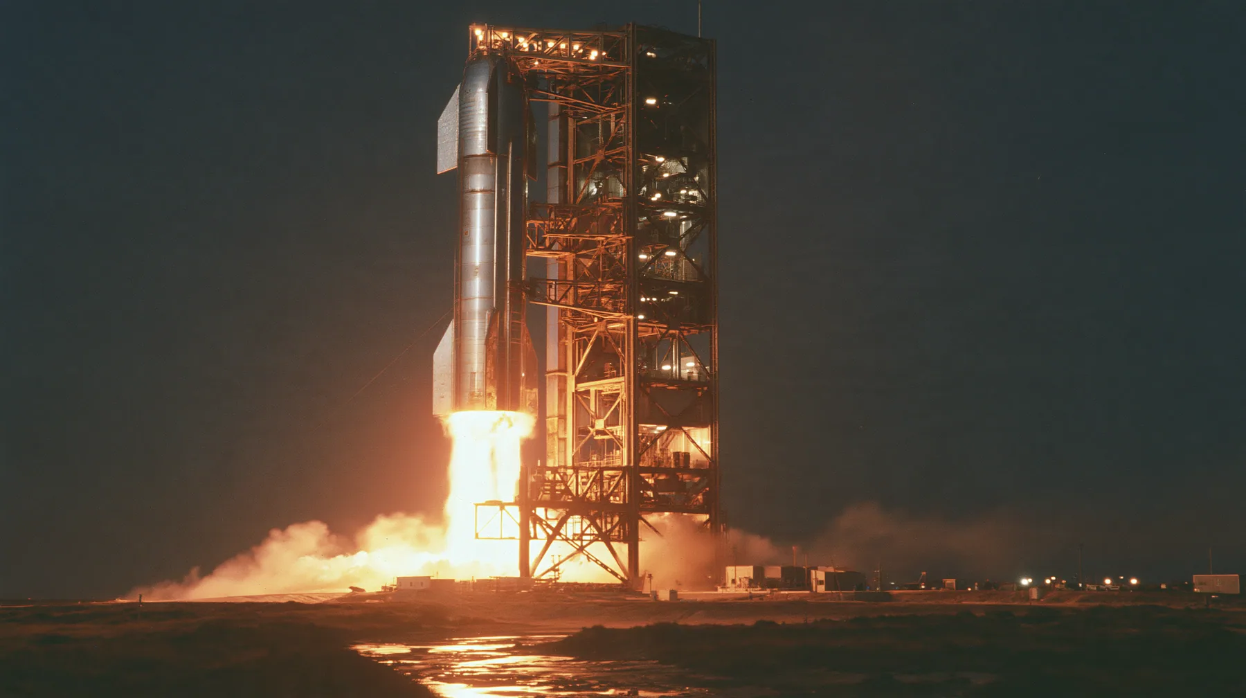 a stainless-steel rocket booster descending on a column of fire beside a launch tower at night, long exposure, documentary photograph, no text, no letters, no logos