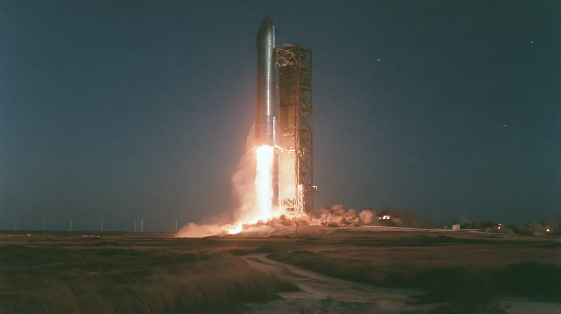 a stainless-steel rocket booster descending on a column of fire beside a launch tower at night, long exposure, documentary photograph, no text, no letters, no logos