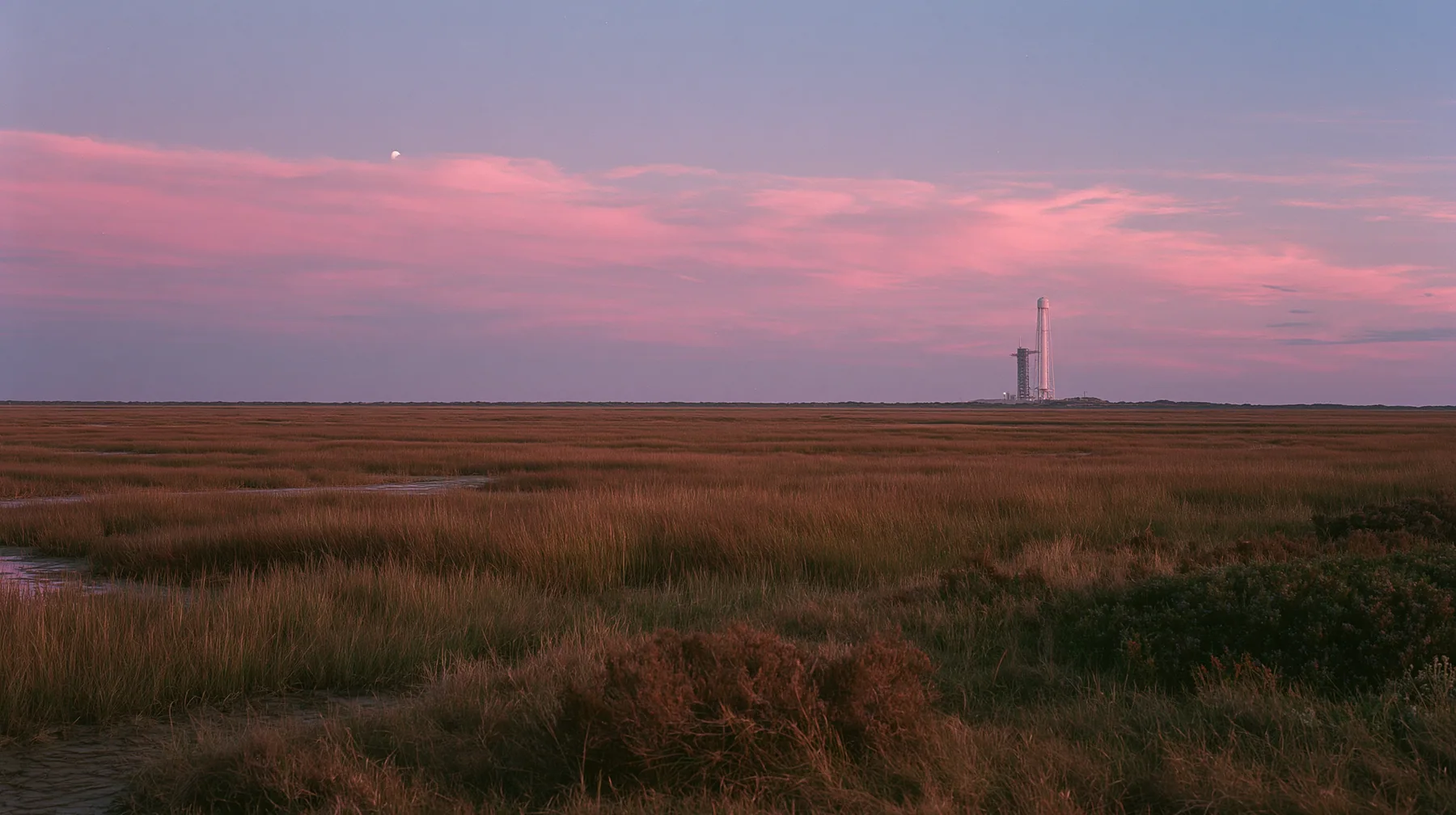 a vast flat Gulf Coast horizon in south Texas at dusk, salt marsh, a single distant launch tower, an enormous pink and violet sky, documentary photograph, no text, no letters, no logos