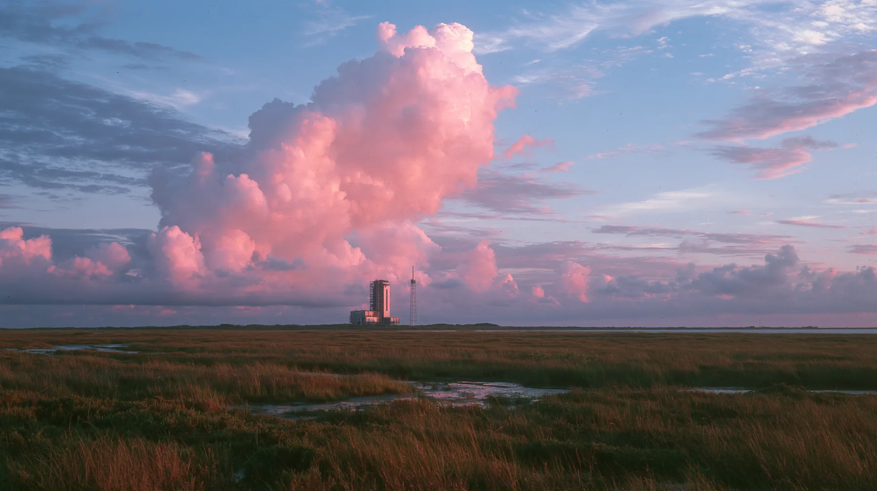 a vast flat Gulf Coast horizon in south Texas at dusk, salt marsh, a single distant launch tower, an enormous pink and violet sky, documentary photograph, no text, no letters, no logos