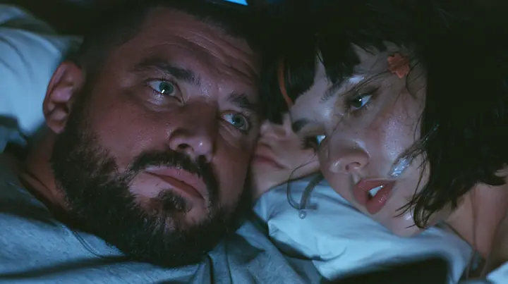 close-up of a broad-shouldered Caucasian man in his early thirties with a square jaw, a short dark beard, a dark buzz cut and heavy brows, the build of a man who lifts, in a grey t-shirt lying in bed in the dark,…