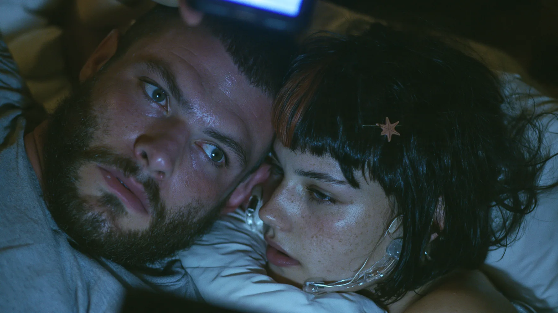 close-up of a broad-shouldered Caucasian man in his early thirties with a square jaw, a short dark beard, a dark buzz cut and heavy brows, the build of a man who lifts, in a grey t-shirt lying in bed in the dark,…