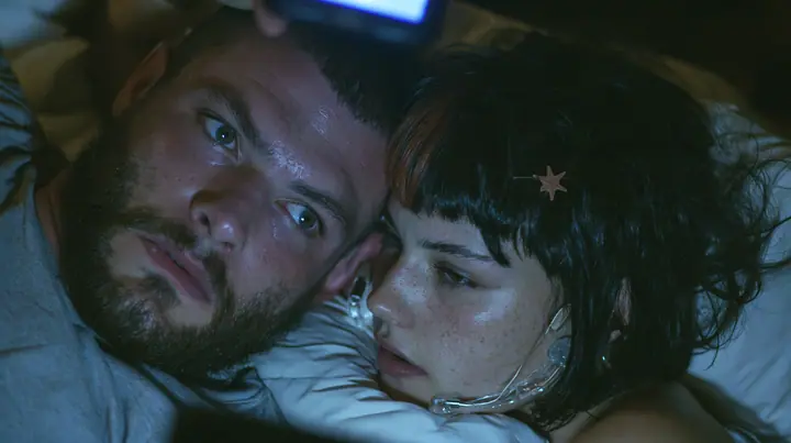 close-up of a broad-shouldered Caucasian man in his early thirties with a square jaw, a short dark beard, a dark buzz cut and heavy brows, the build of a man who lifts, in a grey t-shirt lying in bed in the dark,…