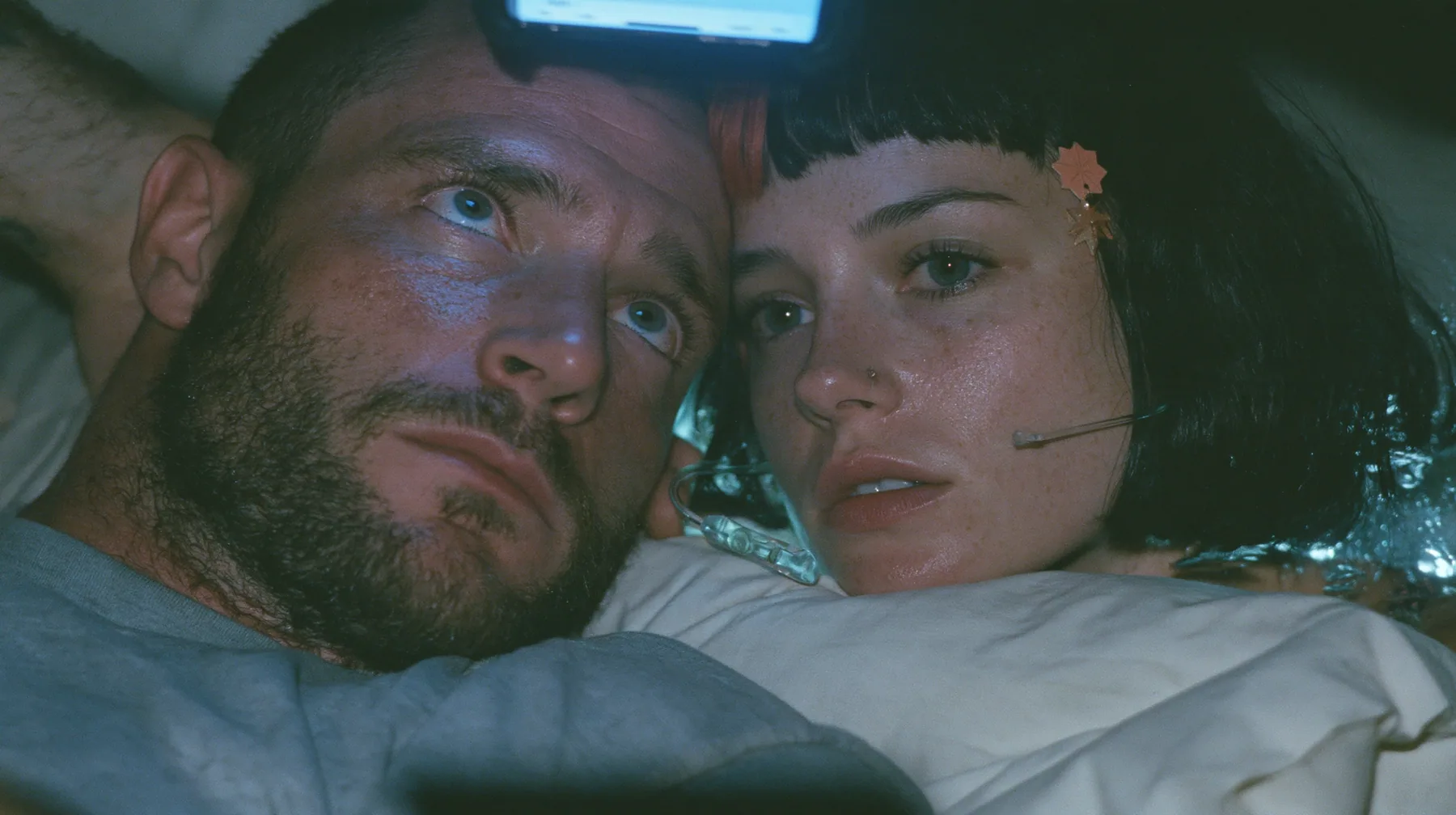 close-up of a broad-shouldered Caucasian man in his early thirties with a square jaw, a short dark beard, a dark buzz cut and heavy brows, the build of a man who lifts, in a grey t-shirt lying in bed in the dark,…