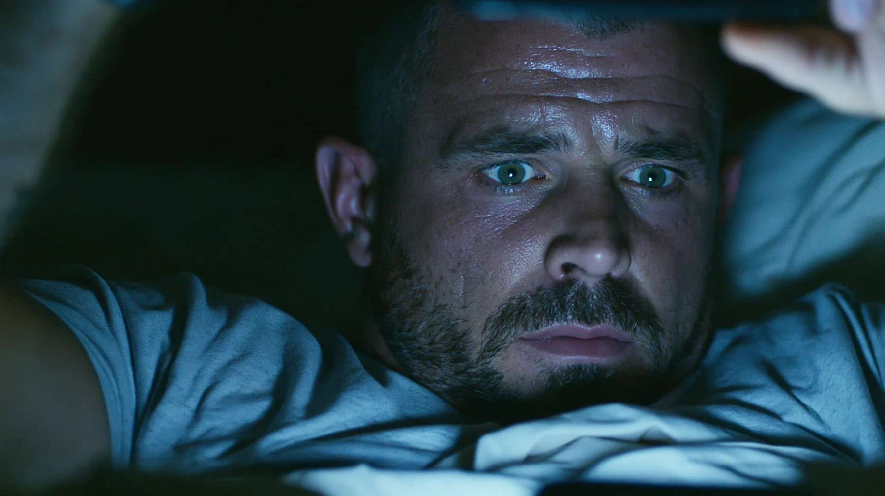 close-up of a broad-shouldered Caucasian man in his early thirties with a square jaw, a short dark beard, a dark buzz cut and heavy brows, the build of a man who lifts, in a grey t-shirt lying in bed in the dark,…