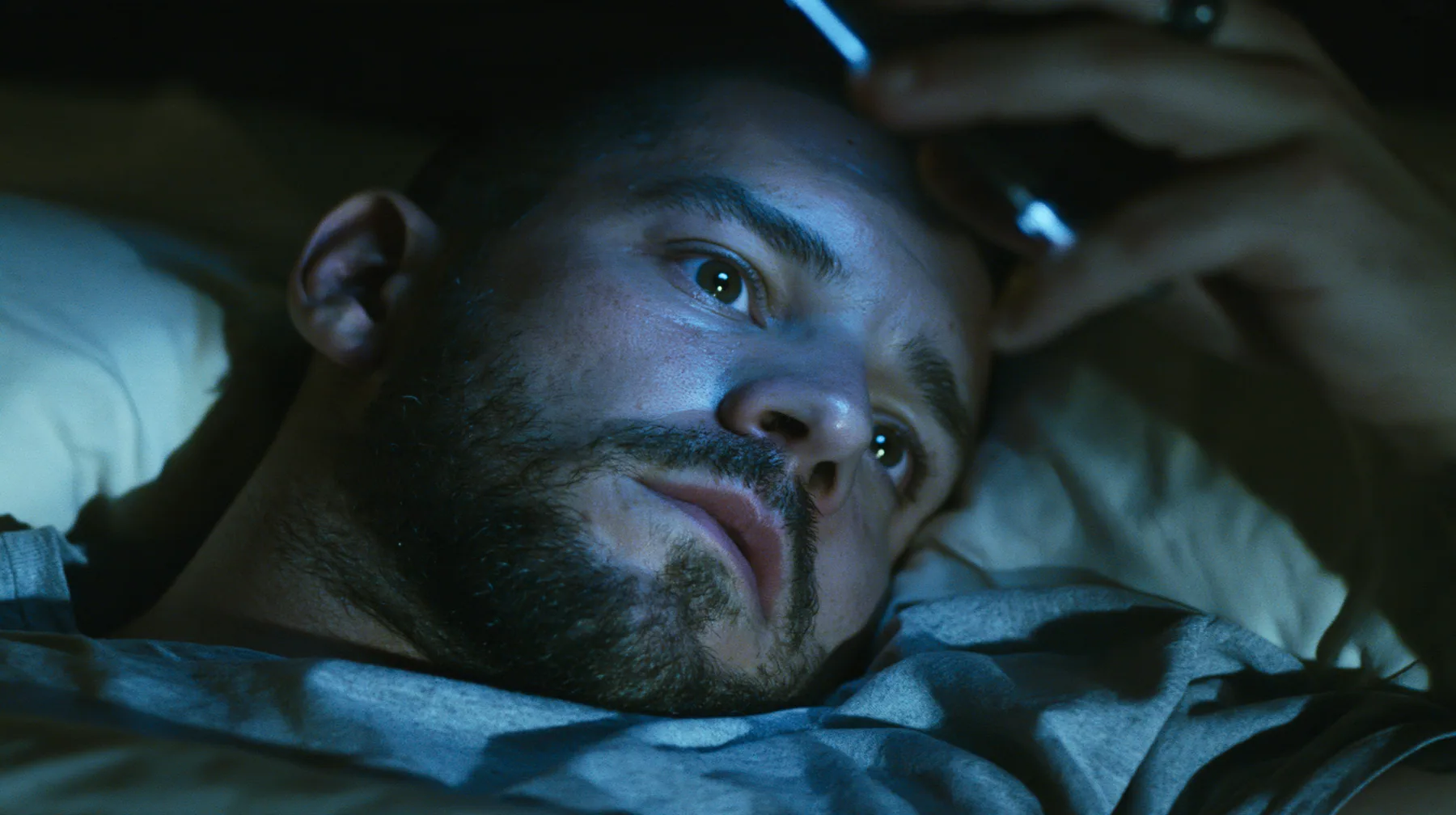 close-up of a broad-shouldered Caucasian man in his early thirties with a square jaw, a short dark beard, a dark buzz cut and heavy brows, the build of a man who lifts, in a grey t-shirt lying in bed in the dark,…