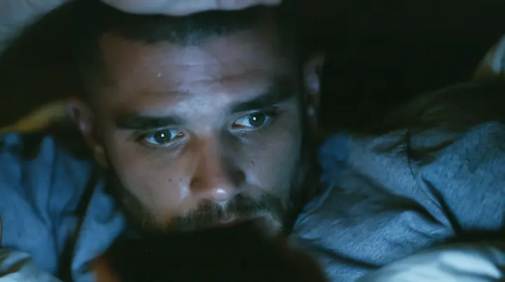 close-up of a broad-shouldered Caucasian man in his early thirties with a square jaw, a short dark beard, a dark buzz cut and heavy brows, the build of a man who lifts, in a grey t-shirt lying in bed in the dark,…