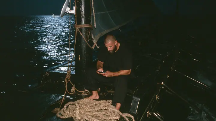 medium wide shot of a broad-shouldered Caucasian man in his early thirties with a square jaw, a short dark beard, a dark buzz cut and heavy brows, the build of a man who lifts, in a plain black t-shirt and black…