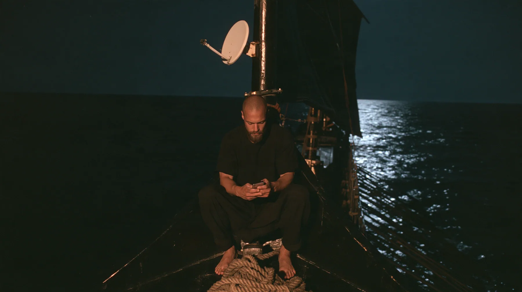 medium wide shot of a broad-shouldered Caucasian man in his early thirties with a square jaw, a short dark beard, a dark buzz cut and heavy brows, the build of a man who lifts, in a plain black t-shirt and black…