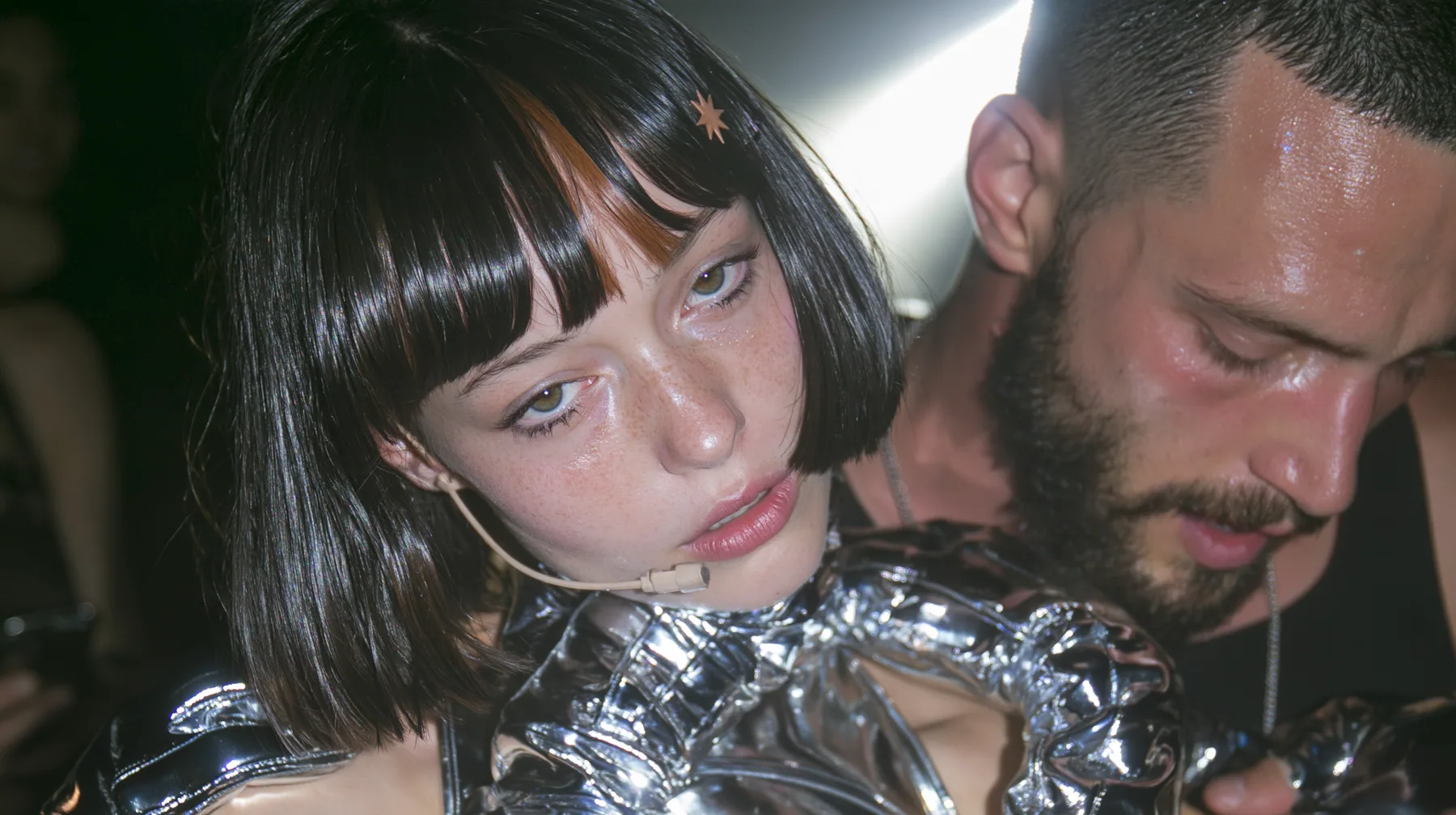 medium close-up of a Caucasian American woman in her mid-twenties with pale skin and light freckles, a glossy black blunt jaw-length bob with heavy straight bangs and one clay-orange streak through the bangs, a small…