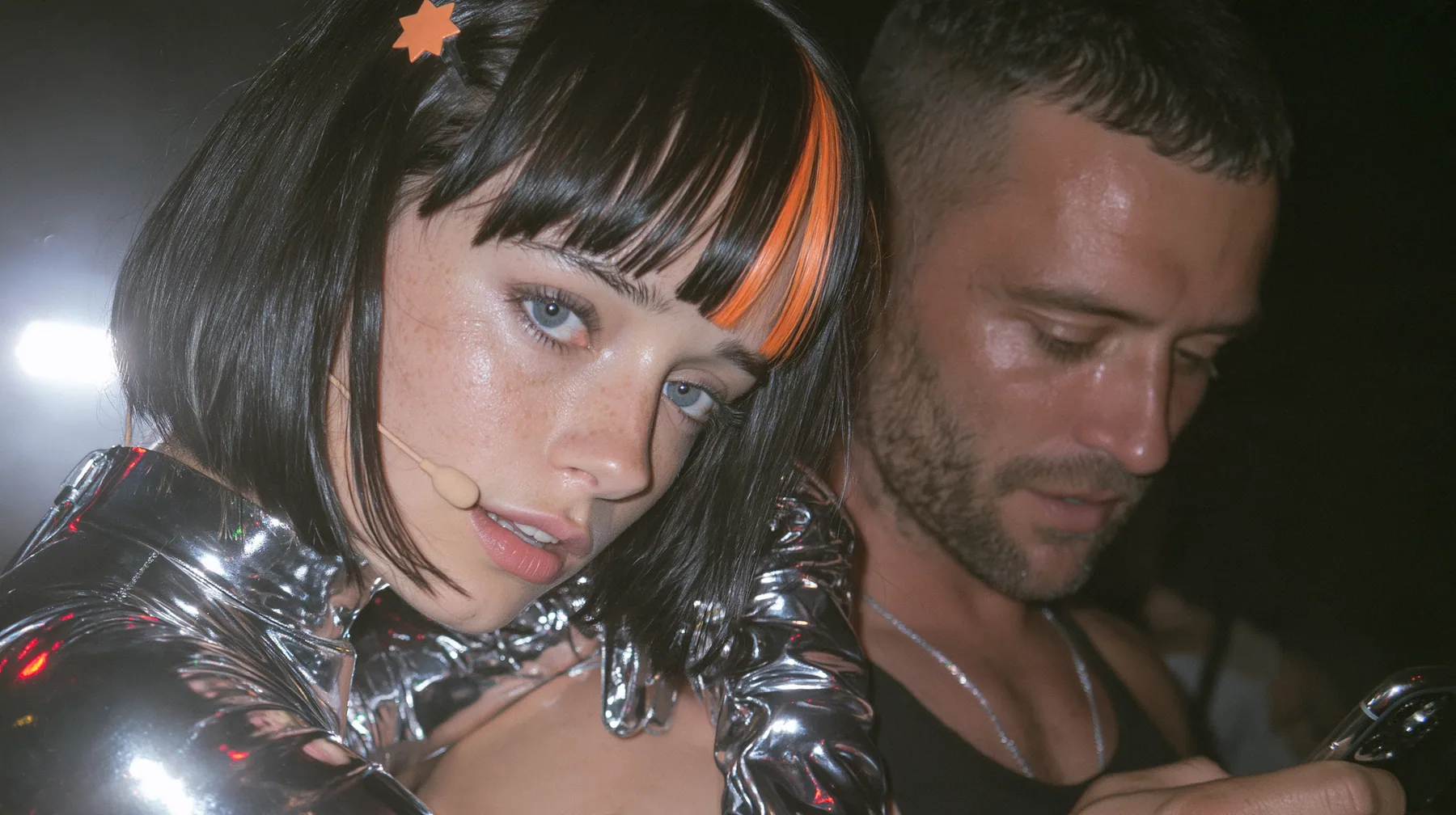 medium close-up of a Caucasian American woman in her mid-twenties with pale skin and light freckles, a glossy black blunt jaw-length bob with heavy straight bangs and one clay-orange streak through the bangs, a small…