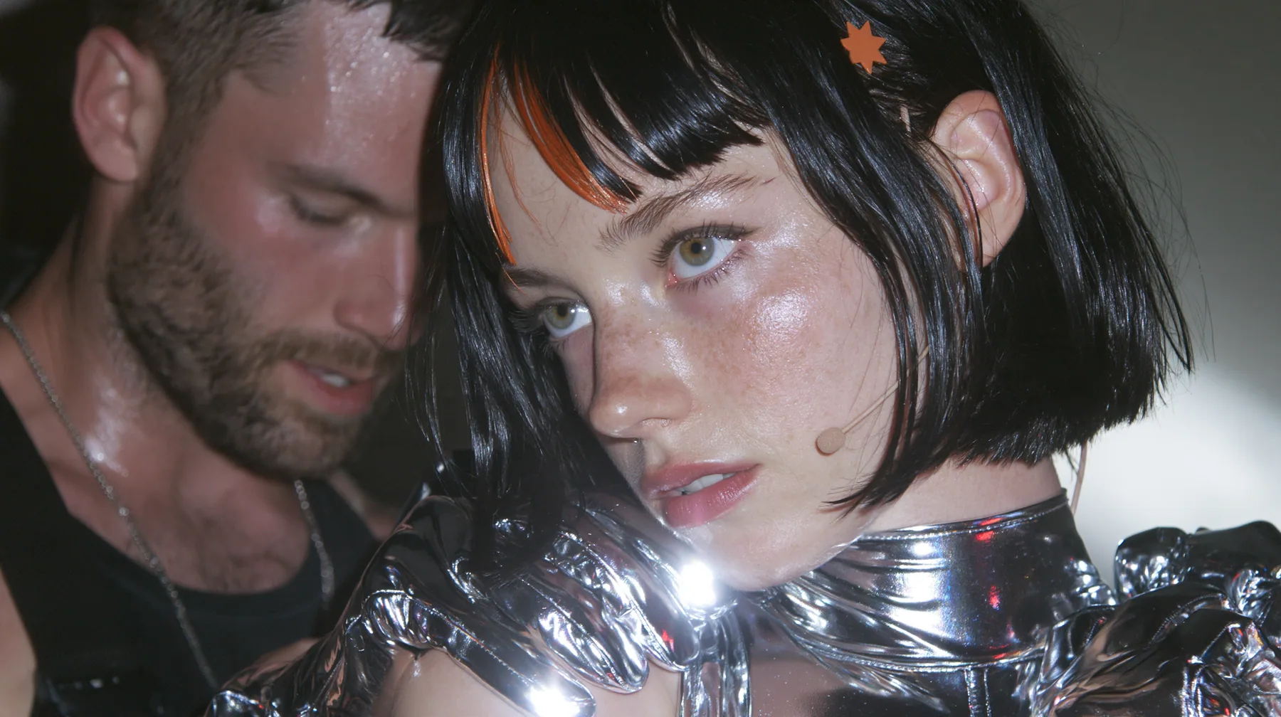 medium close-up of a Caucasian American woman in her mid-twenties with pale skin and light freckles, a glossy black blunt jaw-length bob with heavy straight bangs and one clay-orange streak through the bangs, a small…