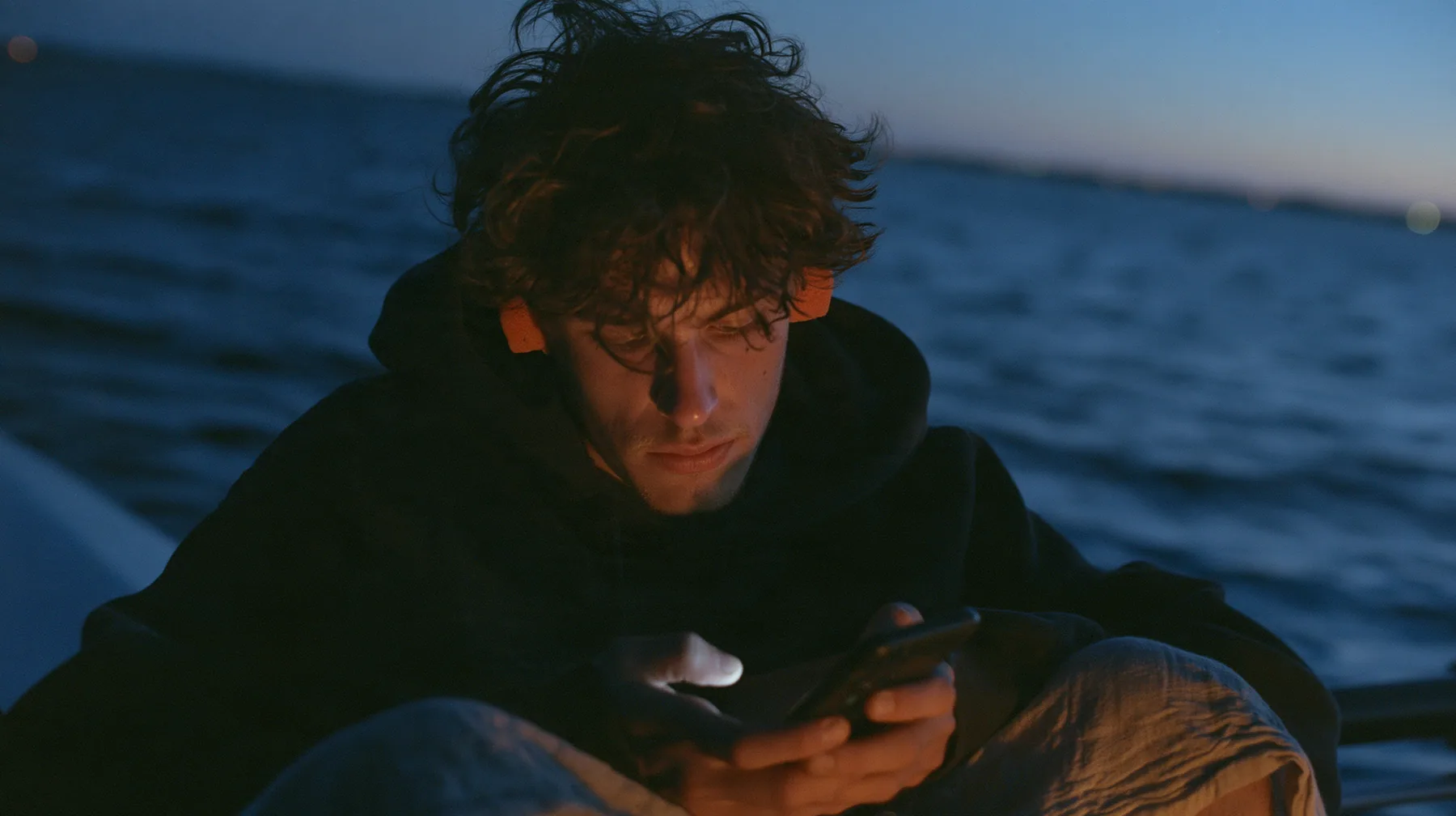 medium close-up of a gaunt young rower in a black hoodie over a linen tunic with orange foam earplugs, looking down at a phone lighting up in his lap, deep blue twilight, the last gold on the horizon, glassy sea,…
