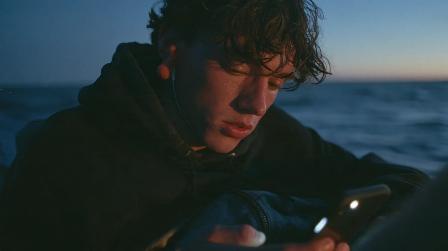 medium close-up of a gaunt young rower in a black hoodie over a linen tunic with orange foam earplugs, looking down at a phone lighting up in his lap, deep blue twilight, the last gold on the horizon, glassy sea,…